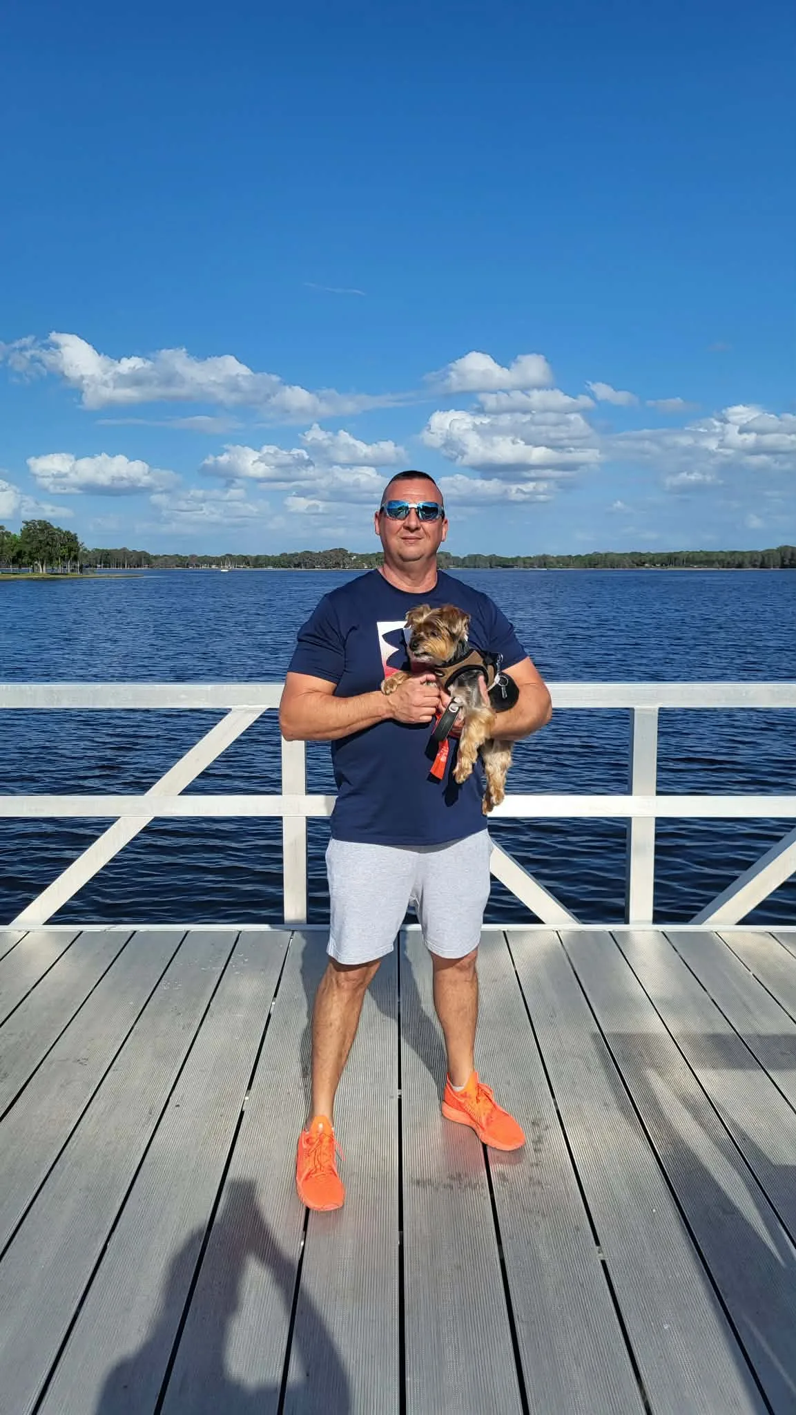 Zsolt Zakara, owner of Silver Painting and Cleaning, holding his small dog Silver on a lakeside dock under a blue sky
