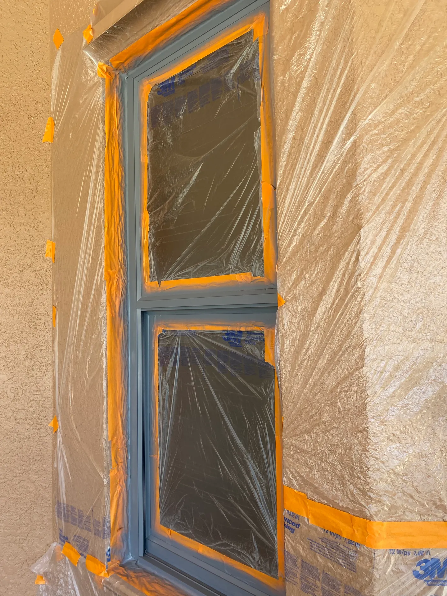 Interior window glass masked with plastic sheeting and orange painter's tape around freshly painted gray window trim, prepped before painting